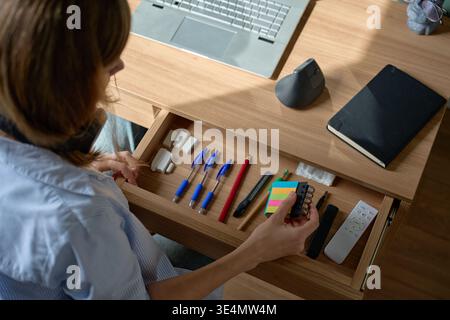 Woman holding sticky note pad over open desk drawer with office supplies near laptop. Concept of workspace organization and home office routine Stock Photo