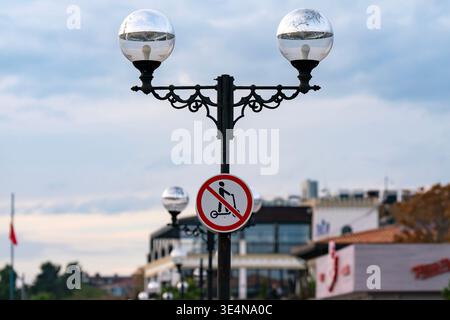 A decorative vintage street lamp displays a red no scooters sign on a busy urban promenade, with blurred buildings and soft evening light creating a c Stock Photo