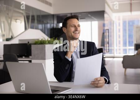 Smiling Hispanic entrepreneur reviewing documents by laptop at office desk Stock Photo
