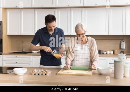 Older woman flattening dough cooking in kitchen with adult son Stock Photo