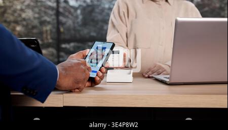 Young Man Using Smartphone To Pay Bill In Busy Cafe Environment. African Man Making Quick Contactless Payment Stock Photo