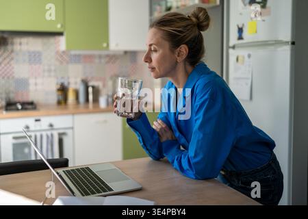 Positive mature woman drinking water and searching for new job vacancies online on laptop. Stock Photo