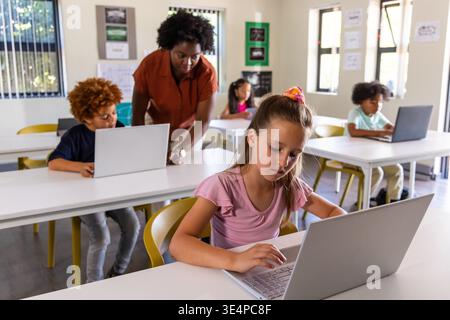 Diverse school-age students with female teacher working on silver laptops at classroom tables Stock Photo