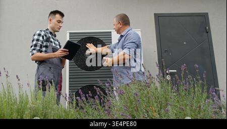 A technician consults with a homeowner at an external heat pump unit, discussing maintenance details. Stock Photo