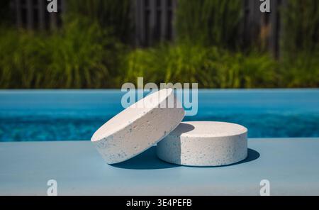 Two large white chlorine tablets with blue specks resting on the poolside edge, used for water disinfection and regular swimming pool maintenance. Stock Photo