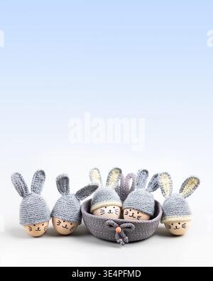Five Easter eggs with funny faces and gray crocheted hats with bunny ears in a decorative basket on a gray blue gradient background. Happy Easter concept. Greeting card. Stock Photo