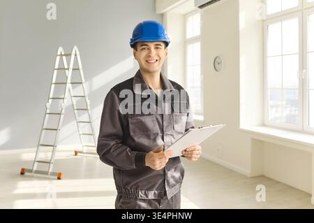 Smiling construction worker in helmet with clipboard in bright interior and ladder Stock Photo