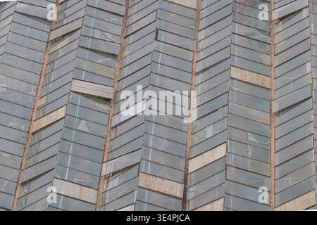 Close-up textured view of an aged industrial greenhouse glass roof featuring a repeating chevron or zigzag pattern of weathered glass panels and metal Stock Photo