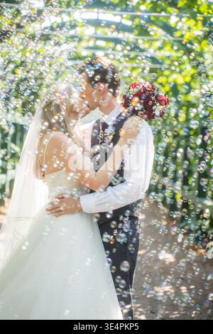 Bride and groom are walking together in a park. They are kissing. Couple is blurred and the soap bubbles are flying in the foreground. Stock Photo