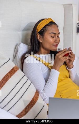Asian woman holding glass cup near face, reclining on bed with mustard pinafore and laptop Stock Photo