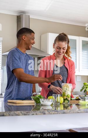 Diverse couple preparing green smoothie at kitchen island using blender jar, apples, yogurt Stock Photo
