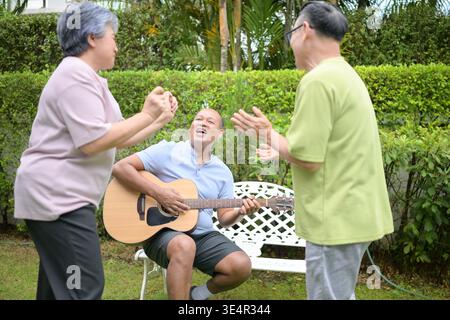 Group of asian seniors singing and dancing while their friend plays guitar in a garden, enjoying friendship and outdoor leisure activity || Model released Stock Photo