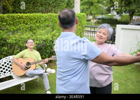 Group of asian seniors singing and dancing while their friend plays guitar in a garden, enjoying friendship and outdoor leisure activity || Model released Stock Photo