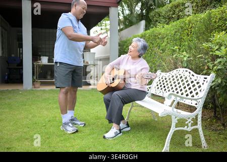 Senior woman sitting on a bench in the garden playing an acoustic guitar while a friend teaching. Concept of music learning and creative leisure in retirement || Model released Stock Photo
