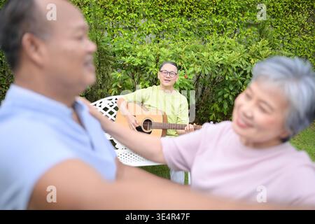 Group of asian seniors singing and dancing while their friend plays guitar in a garden, enjoying friendship and outdoor leisure activity || Model released Stock Photo
