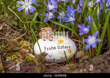 München, Deutschland 28.03.2026: Im Bild: Dekoratives Osterei mit der Aufschrift Frohe Ostern und kleiner Hasenfigur liegt zwischen blühenden Frühlingsblumen in einem Gartenbeet. Die Szene zeigt typische Osterdekoration im Außenbereich und verbindet florale Motive mit saisonaler Gestaltung im Frühling. Bayern *** Munich, Germany 28 03 2026 In the picture Decorative Easter egg with the inscription Happy Easter and small rabbit figure lies between blooming spring flowers in a garden bed The scene shows typical Easter decoration in the outdoor area and combines floral motifs with seasonal design Stock Photo