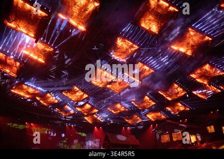 Fiery glow illuminates dark, industrial ceiling structure. Orange lights cast dramatic, angular patterns across metal framework. Smoke drifts subtly t Stock Photo