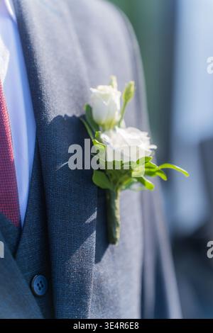 A man in a gray jacket and white shirt. It has a black bowtie on it. In the jacket pocket there is a bouquet of white flowers with green leaves Stock Photo