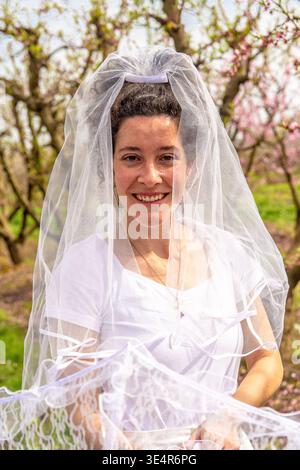 Woman standing in a blooming peach orchard, smiling for the camera while wearing a white wedding veil and simple t shirt, celebrating spring and roman Stock Photo