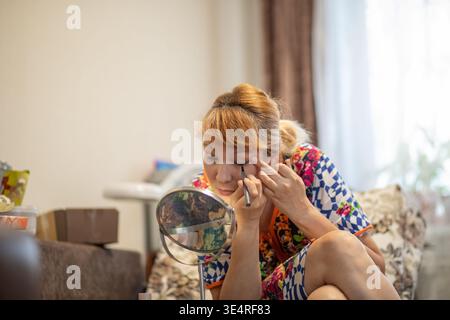 Middle aged woman applying eyeliner looking at small mirror in living room. Daily morning beauty makeup routine of 57 year old lady. Self care and agi Stock Photo