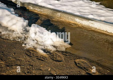 Melting snow with flowing water along curb on muddy road, close up. Concept: seasonal snow melt Stock Photo