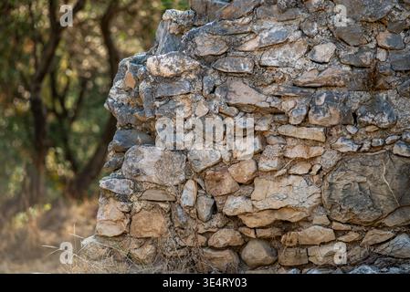 Close-up of a dry-stone wall made of rough limestone rocks, weathered texture in warm light with blurred woodland background. Stock Photo