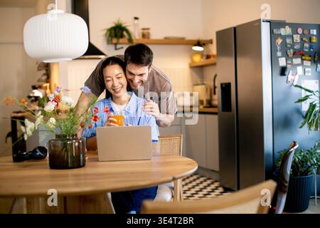 A man and a woman share a joyful moment in a kitchen. The woman works on a laptop while the man stands behind her, smiling. Flowers are on the table a Stock Photo