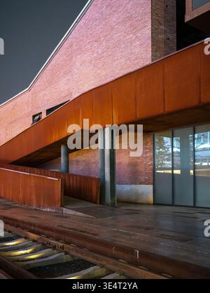 A diagonal rusty corten steel architectural ramp crossing a renovated red brick building facade above a wooden deck at night. The scene represents ind Stock Photo