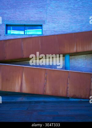 A diagonal rusty corten steel ramp structure crossing a red brick building facade illuminated by vibrant blue lighting at night. The scene represents Stock Photo
