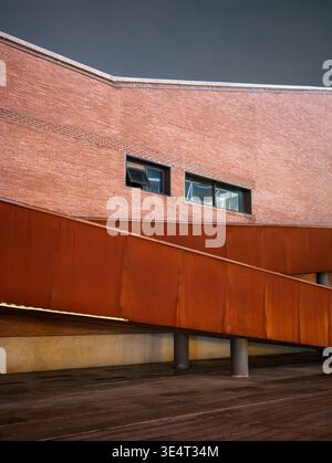 A diagonal rusty corten steel architectural structure intersecting a renovated red brick building facade with a small open window at night. The scene Stock Photo