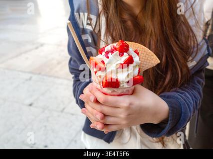 Young woman enjoys a colorful ice cream sundae with strawberries and whipped cream, holding it in both hands while standing outdoors in an urban area Stock Photo
