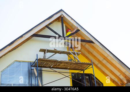 Professional in yellow shirt seals joint in decorative wooden trim, house gable exterior, working elevated, visible caulk gun and safety helmet. Stock Photo