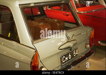 Rear view of a light blue Moskvich van with a Soviet license plate, showing two brown suitcases through the back window and part of a red car. Stock Photo