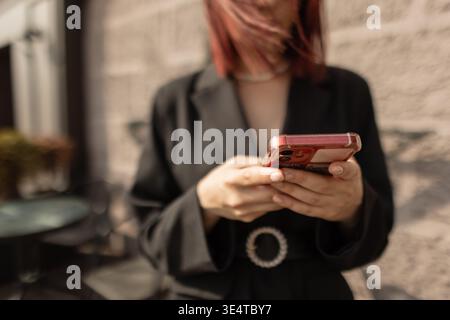 Close-up of young woman with smartphone, city background blurred. Focus on hands, smart casual style, sunny day. Attractive woman in blazer reading me Stock Photo
