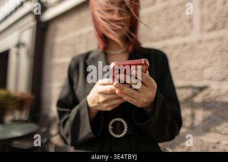 Stylish lady in eyeglasses checking phone on sunny street. Business casual fashion, city background, modern tech-savvy woman. Stock Photo