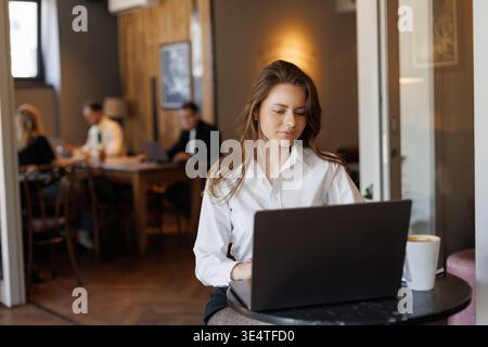 Young woman working on a laptop in a cozy cafe. She wears a white blouse and polka dot tights, sitting at a small round table with coffee and a smartp Stock Photo