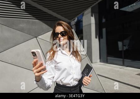 Stylish young woman in business outfit checking her phone outside. Professional, tech-savvy, and independent in a vibrant cityscape. Stock Photo