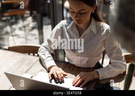 Woman using laptop, sitting by window with sunlight casting shadows. Freelance and tech concept. Elegant female worker focused on task, typing on lapt Stock Photo