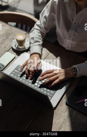 Freelancer or remote worker using laptop at wooden desk. Daylight, coffee, and gadgets create a modern work-from-anywhere atmosphere. Stock Photo