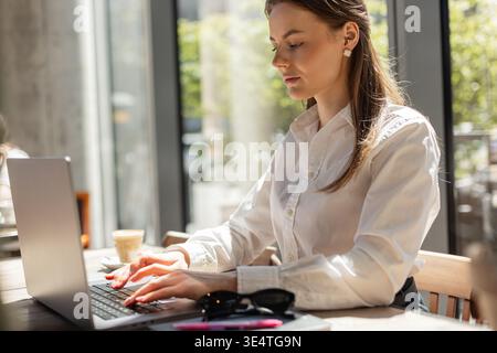 Freelancer woman with coffee cup working on laptop by the window. Business and lifestyle concept. Confident businesswoman working on laptop at modern Stock Photo