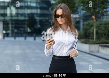 Professional woman using smartphone, holding notebook, dressed in formal attire. Confident and focused in a city business district. Businesswoman in s Stock Photo