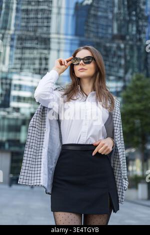 Confident young businesswoman in stylish outfit and sunglasses standing in modern city center. Professional fashion and urban lifestyle concept. Moder Stock Photo