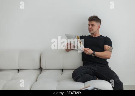 Stylish handsome fashion business man in black clothes reading magazine and relaxing on white sofa in room. Cool guy rest Stock Photo