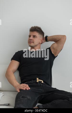 Stylish handsome muscular man in fashion black clothes with a T-shirt sits and relaxes on the sofa. Cool business guy Stock Photo