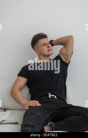 Stylish handsome successful man with hairstyle in fashionable black clothes sits and relaxes on the sofa at home. Cool guy Stock Photo