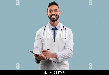 Happy Middle Eastern Doctor Man Taking Notes Standing Over Blue Background Stock Photo
