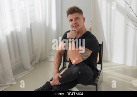 Handsome fashionable successful man in black t-shirt sitting on chair in room. Business man Stock Photo