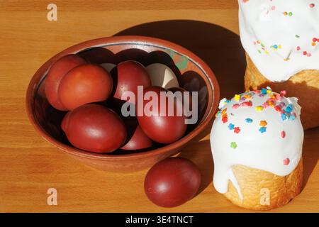 Traditional Easter red eggs in a clay bowl with festive glazed cake on wooden table, rustic style with copy space. Stock Photo