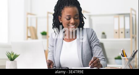 Black Millennial Businesswoman Taking Notes And Working On Laptop In Office Stock Photo