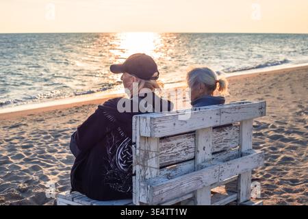 A man and a old woman sit on a wooden bench on a sandy beach looking at the sunset over the water. father and son, beach vacation, sunset view. Stock Photo